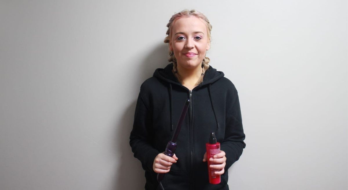 A female holding curling tongs and a spray bottle in front of a white wall